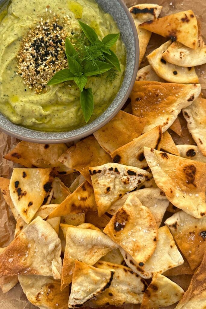 Zucchini yogurt dip topped with dukkah, olive oil and fresh herbs and served in a grey stoneware bowl. The bowl of zucchini yogurt dip is surrounded by sourdough pita chips.