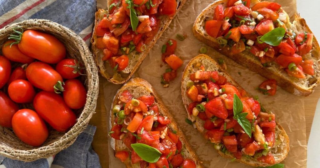 4 slices of sourdough bruschetta sitting on a piece of parchment paper. There is a basket of Roma tomatoes sitting to the left of the photo.
