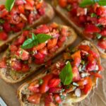 4 slices of sourdough bruschetta sitting on a piece of parchment paper. There is a basket of Roma tomatoes sitting to the left of the photo.