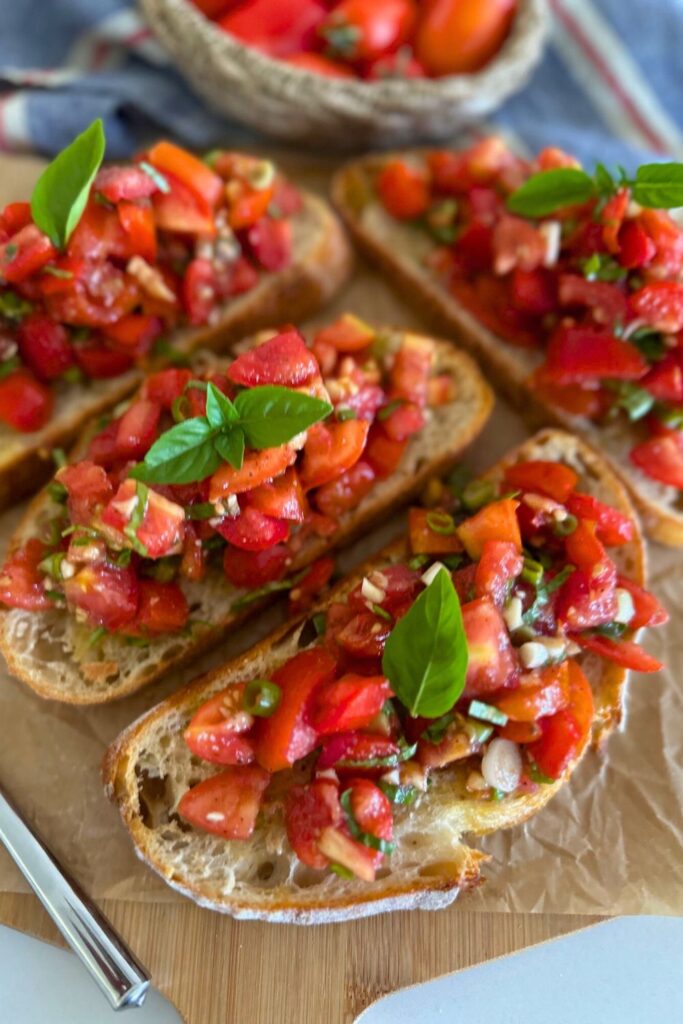 4 slices of sourdough bruschetta sitting on a piece of parchment paper. There is a basket of Roma tomatoes sitting to the left of the photo.