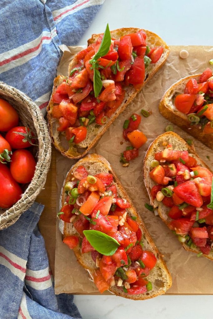 4 slices of sourdough bruschetta sitting on a piece of parchment paper. There is a basket of Roma tomatoes sitting to the left of the photo.
