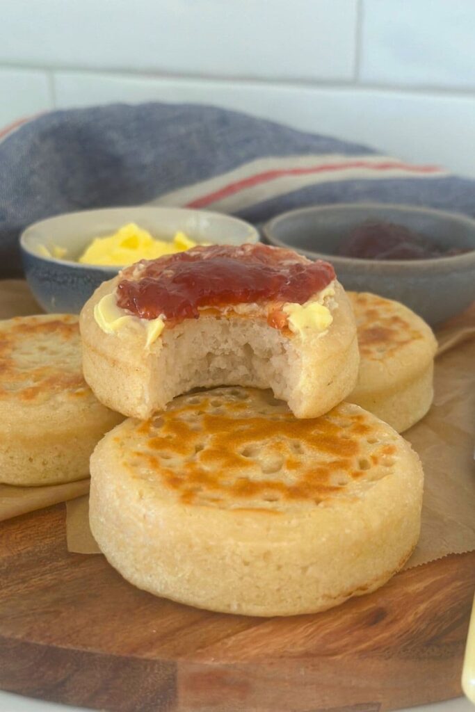 A selection of sourdough crumpets sitting on a wooden board. The sourdough crumpet at the top of the pile is smothered in strawberry jam and has a bite taken out of it.