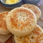 4 sourdough crumpets displayed on a wooden board. There are 2 bowls of butter and jam sitting behind the sourdough crumpets.