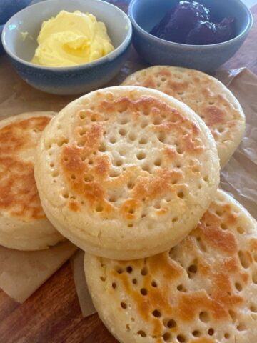 4 sourdough crumpets displayed on a wooden board. There are 2 bowls of butter and jam sitting behind the sourdough crumpets.