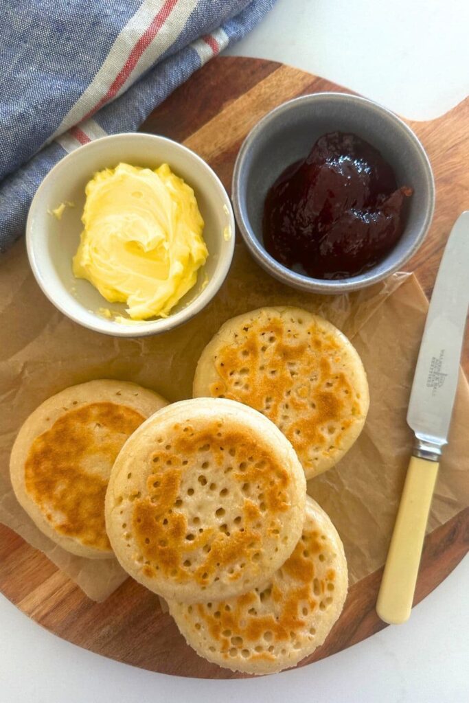 4 sourdough crumpets sitting on a wooden board with a bowl of jam and butter next to an ivory handled knife.