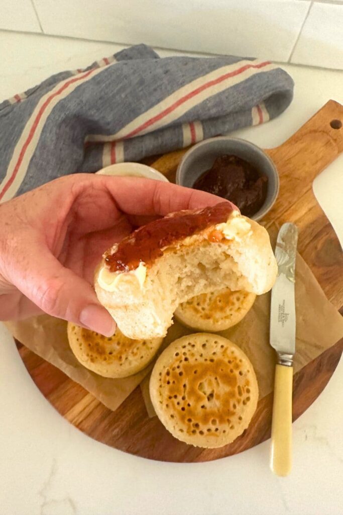 A selection of sourdough crumpets sitting on a wooden board. The sourdough crumpet at the top of the pile is smothered in strawberry jam and has a bite taken out of it.