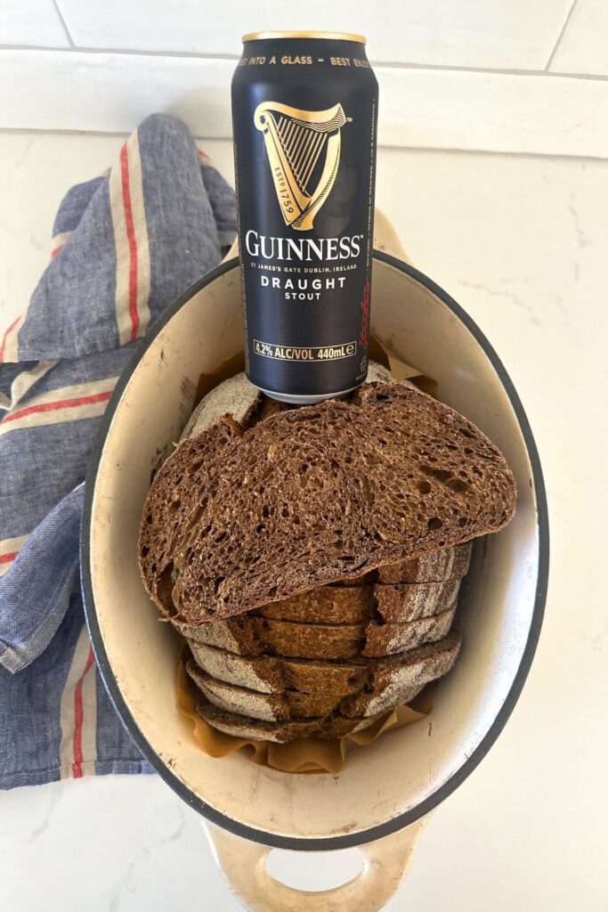 A loaf of sourdough bread that has been made with Guinness. The dark loaf of sourdough is sitting in a cream enamel Dutch Oven and there is a can of Guinness sitting on the edge of the Dutch Oven.