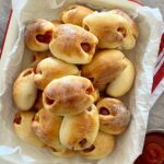 A dish of sourdough pigs in blankets ready for sourdough super bowl snacking. The cream enamel dish is sitting on a red striped dish towel. There is a bowl of tomato ketchup to the right of the sourdough pigs in blankets.