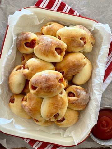 A dish of sourdough pigs in blankets ready for sourdough super bowl snacking. The cream enamel dish is sitting on a red striped dish towel. There is a bowl of tomato ketchup to the right of the sourdough pigs in blankets.