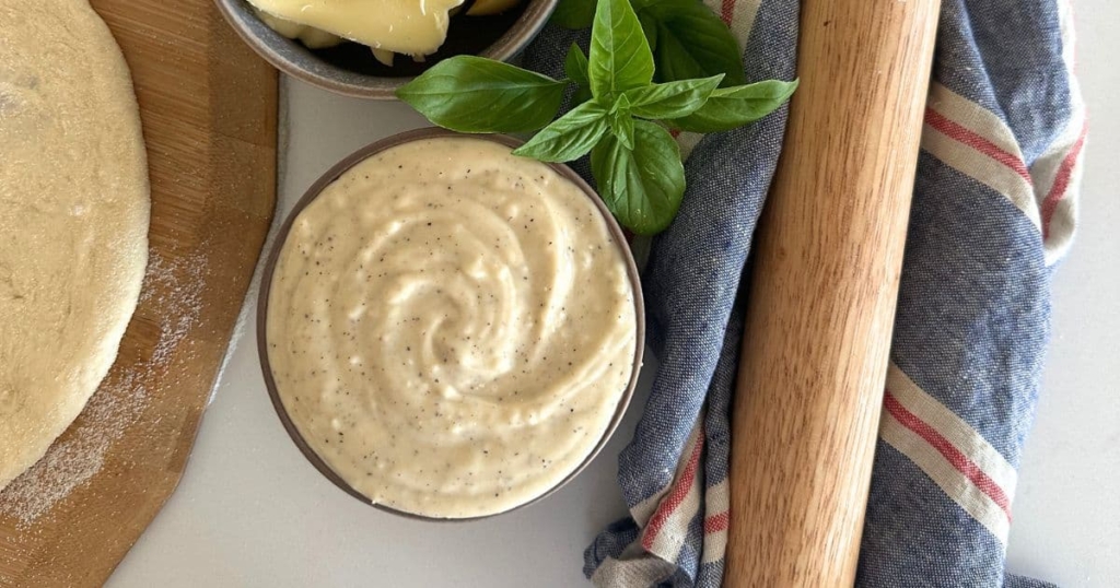 A bowl of white pizza sauce made with sourdough starter, sitting on a white counter top next to a pizza base, some basil leaves and a rolling pin.