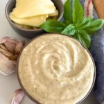 A bowl of white pizza sauce made with sourdough starter sitting on a white counter top. The bowl is surrounded by garlic cloves, cheese and basil leaves.