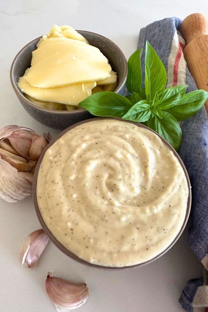 A bowl of white pizza sauce made with sourdough starter sitting on a white counter top. The bowl is surrounded by garlic cloves, cheese and basil leaves.
