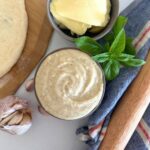 A bowl of white pizza sauce made with sourdough starter sitting on a white counter top. The bowl is surrounded by garlic cloves, cheese and basil leaves.