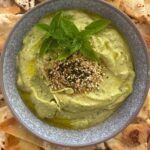 Zucchini yogurt dip topped with dukkah, olive oil and fresh herbs and served in a grey stoneware bowl. The bowl of zucchini yogurt dip is surrounded by sourdough pita chips.