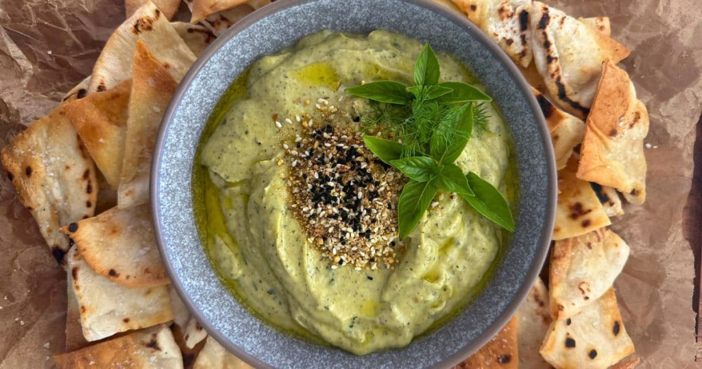 ZUCCHINI YOGURT DIP served in a grey stoneware bowl. The dip is topped with olive oil, dukkah and fresh herbs. The bowl is surrounded with sourdough pita chips.