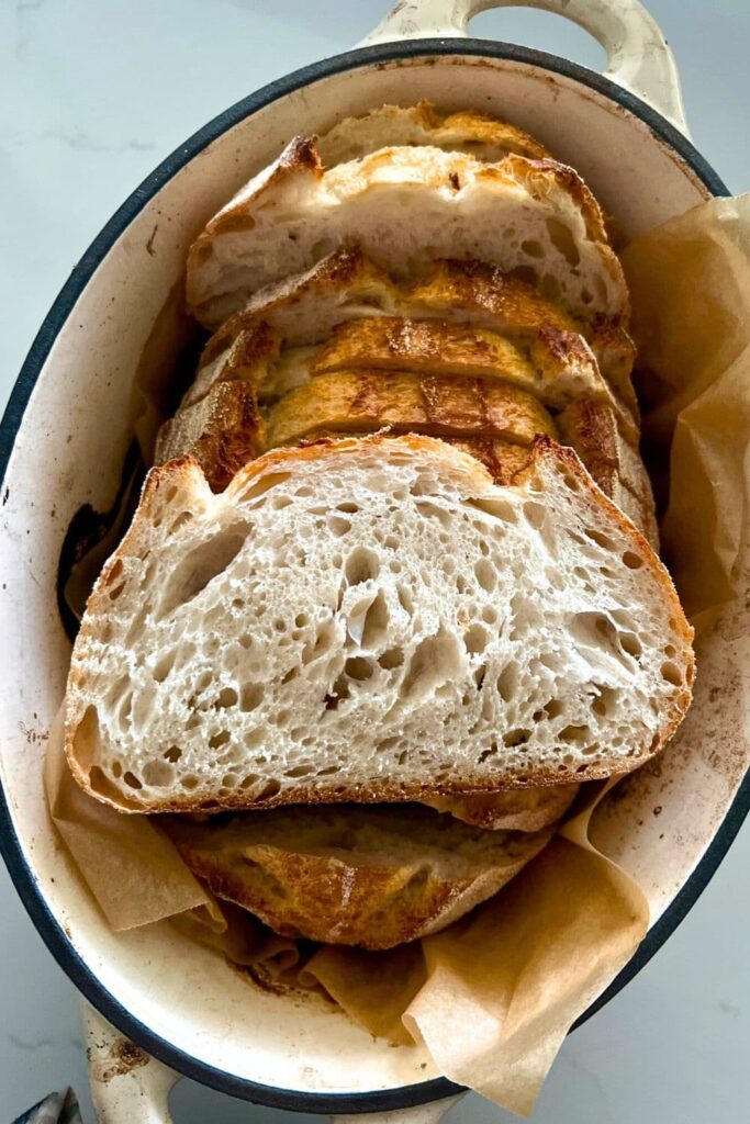 A loaf of sourdough bread that has been sliced up and then put back into the Dutch Oven. A slice has been layed on top of the loaf so you can see the light, airy crumb.