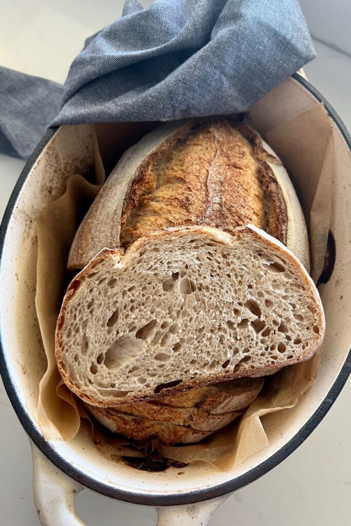 A loaf of spelt sourdough bread sitting in a cream enamel Dutch Oven. There is a slice sitting on the top to show the crumb of the loaf.