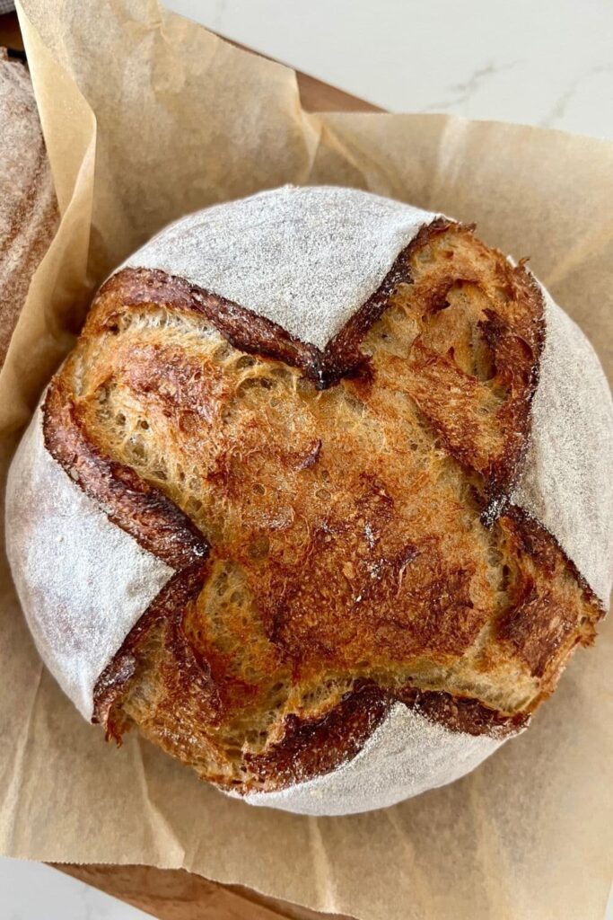 A round loaf of sourdough rye bread that has been scored with a cross. It has been dusted in white rice flour and sits on a sheet of brown parchment paper. 