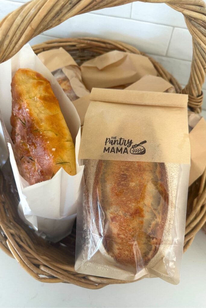 A cane basket filled with small sourdough loaves in brown paper bags. There is a also a slab of sourdough focaccia in a white paper bag.