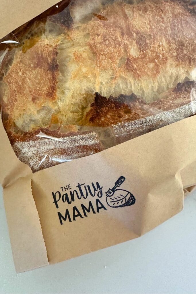 A close up image of a brown paper and cellophane bread bag that has been stamped with "The Pantry Mama" logo stamp. There is a crusty loaf of sourdough bread visible through the cellophane window of the bread bag.
