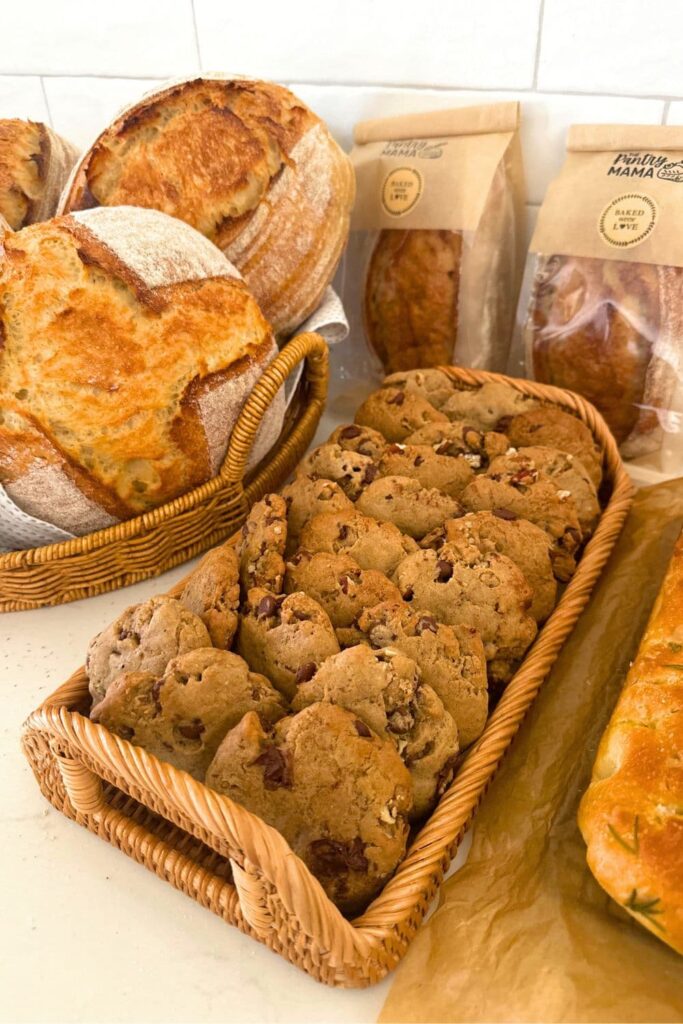 A tray of sourdough chocolate chip cookies nestled among loaves of sourdough bread ready to be sold from a home sourdough bakery.