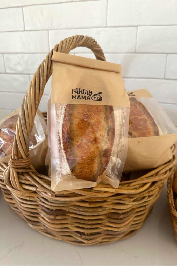 A large cane basket filled with small sourdough loaves packaged in brown paper and cellophane bread bags.