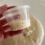 Someone holding a small aliquot jar over a glass bowl of sourdough.