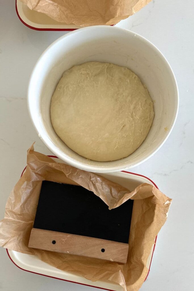 A cream bowl filled with sourdough focaccia dough. There is also a red edged enamel tray lined with parchment paper.