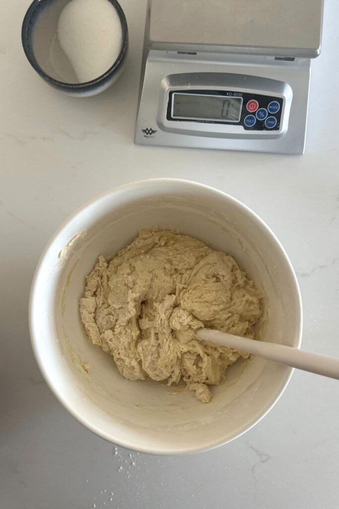 A cream mixing bowl filled with sourdough focaccia dough. You can see a digital scale in the top of the photo.