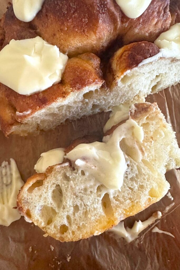 Close up photo of sourdough cinnamon roll focaccia bread. There is a slice turned on its side so you can see the soft interior crumb. The whole loaf is drizzled in creamy vanilla cream cheese frosting.