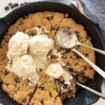 Sourdough Cookie Skillet with chocolate chips - Recipe Feature Image. This image is of a cast iron skillet filled with a giant sourdough chocolate chip cookie baked in the oven and then topped with vanilla ice cream. There are three spoons in the skillet and some of the giant cookie has been eaten.