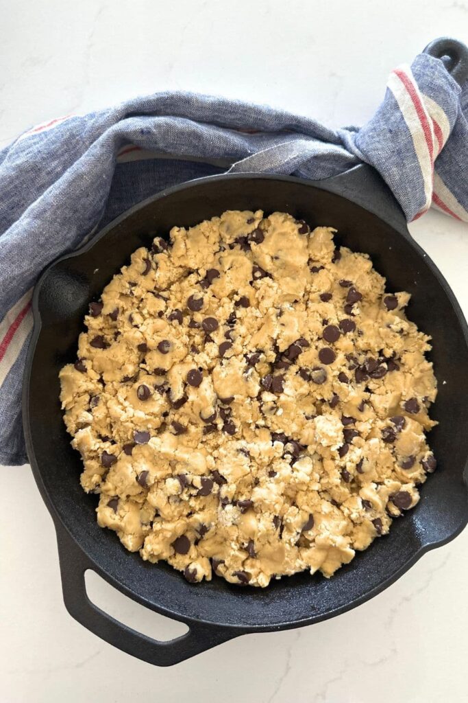 A black cast iron skillet with uncooked sourdough cookie dough in it, ready to go into the oven. There is a blue and white striped dish towel curled around the handle of the cast iron skillet.