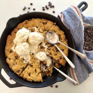 Sourdough Cookie Skillet with chocolate chips - Recipe Feature Image. This image is of a cast iron skillet filled with a giant sourdough chocolate chip cookie baked in the oven and then topped with vanilla ice cream. There are three spoons in the skillet and some of the giant cookie has been eaten.