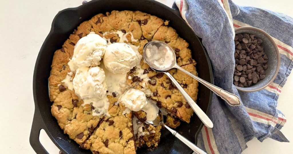 Sourdough Cookie Skillet with chocolate chips. This image is of a cast iron skillet filled with a giant sourdough chocolate chip cookie baked in the oven and then topped with vanilla ice cream. There are three spoons in the skillet and some of the giant cookie has been eaten.