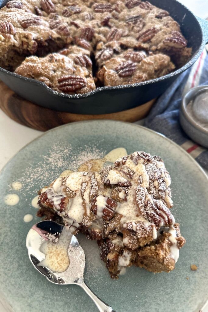 A slice of spiced pecan sourdough cookie skillet drizzled with heavy whipping cream. You can see the cast iron skillet containing the rest of the spiced pecan chocolate chip sourdough skillet cookie in the background.