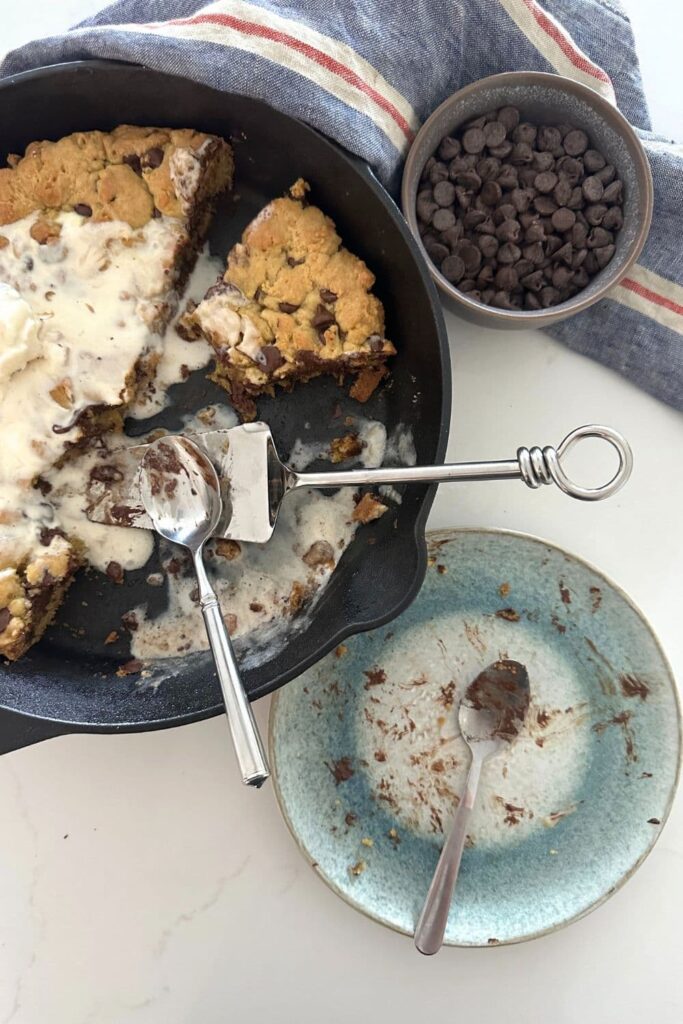 A cast iron skillet containing a half eaten sourdough skillet cookie that was topped with ice cream and cream. There is also a blue speckled dessert plate that has been scraped clean with just a few traces of chocolate chips and ice cream left.