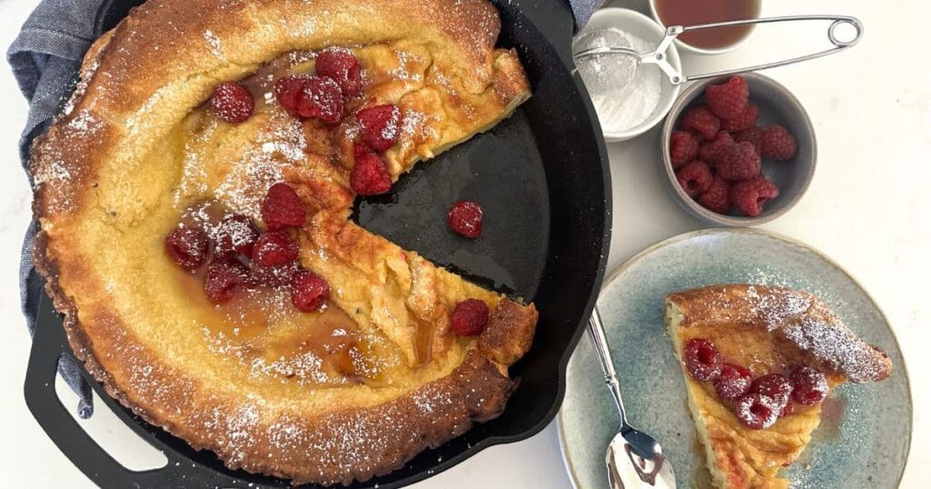 A sourdough Dutch baby pancake served in a cast iron skillet and topped with fresh red raspberries and maple syrup. The German pancake has been dusted with powdered sugar.
