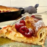 A big slice of sourdough Dutch baby pancake topped with raspberries and maple syrup.