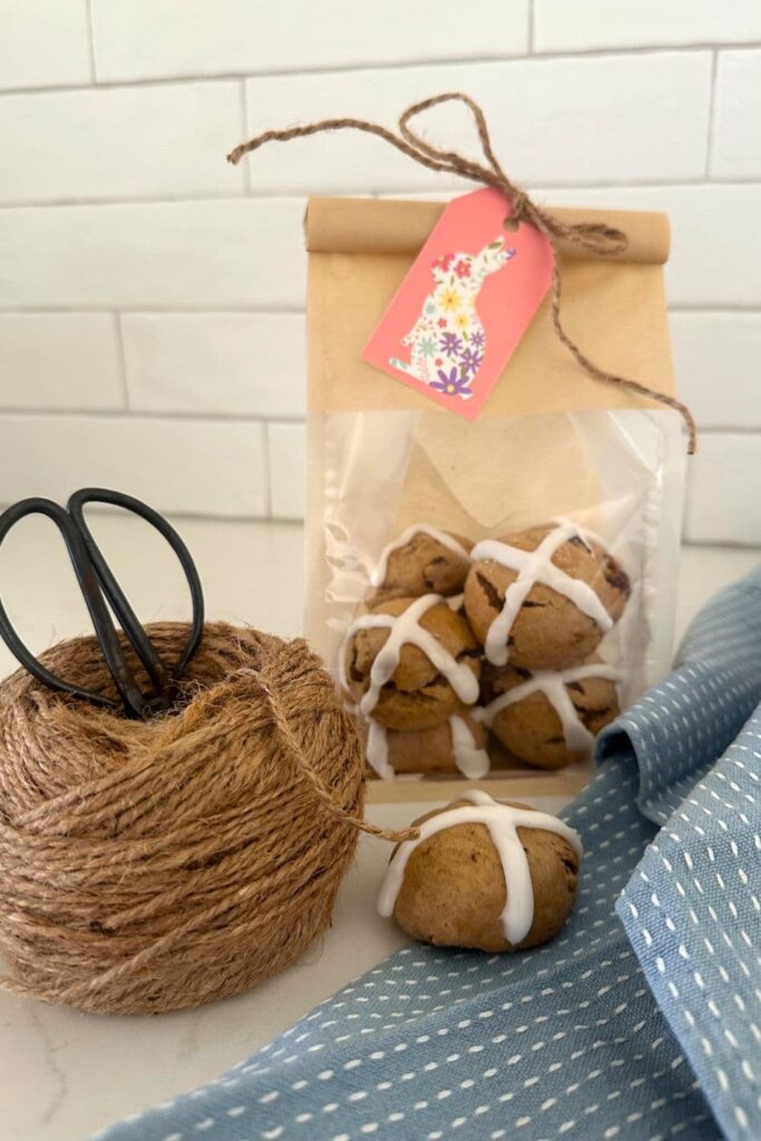 A cellophane gift bag filled with sourdough hot cross cookies. The bag has a pink Easter tag attached. There is a ball of string and antique scissors in the foreground of the photo.