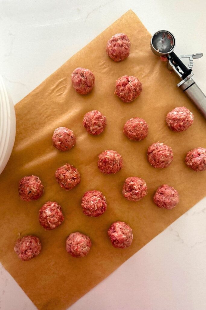 A sheet of parchment paper with sourdough meatballs laid out. There is also a cookie scoop to the right of the meatballs. 