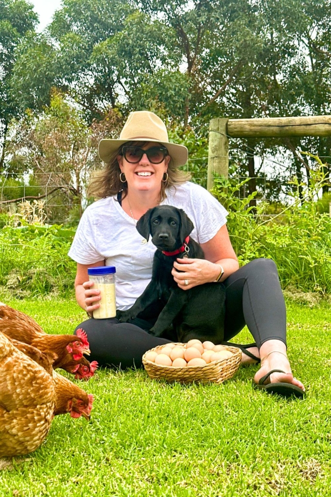 A photo of Kate from The Pantry Mama sitting on a patch of green grass holding a black labrador puppy on her lap. She is holding a jar of sourdough starter with a blue lid and there are two chickens pecking in the grass in front of Kate.