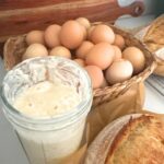 A photo featuring a jar of sourdough starter and a loaf of sourdough bread in front of a basket of fresh eggs.