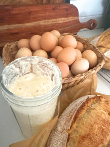 A photo featuring a jar of sourdough starter and a loaf of sourdough bread in front of a basket of fresh eggs.