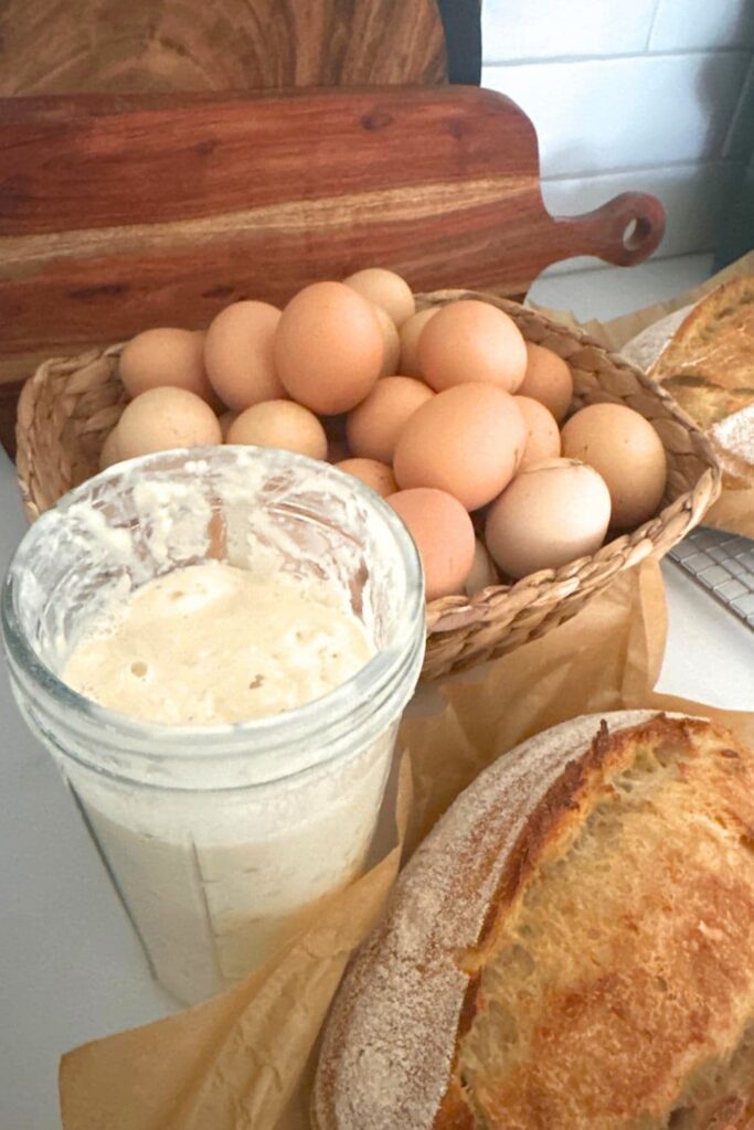 A photo featuring a jar of sourdough starter and a loaf of sourdough bread in front of a basket of fresh eggs. 
