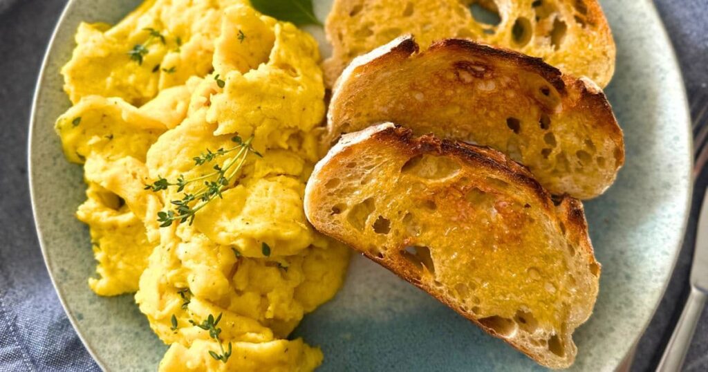 A blue stoneware plate topped with sourdough scrambled eggs, 3 slices of buttered sourdough toast and sitting on a blue dish towel.