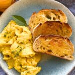 A blue stoneware plate topped with sourdough scrambled eggs, 3 slices of buttered sourdough toast and sitting on a blue dish towel. There is also a glass of orange juice to the left of the plate.