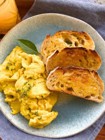 A blue stoneware plate topped with sourdough scrambled eggs, 3 slices of buttered sourdough toast and sitting on a blue dish towel. There is also a glass of orange juice to the left of the plate.