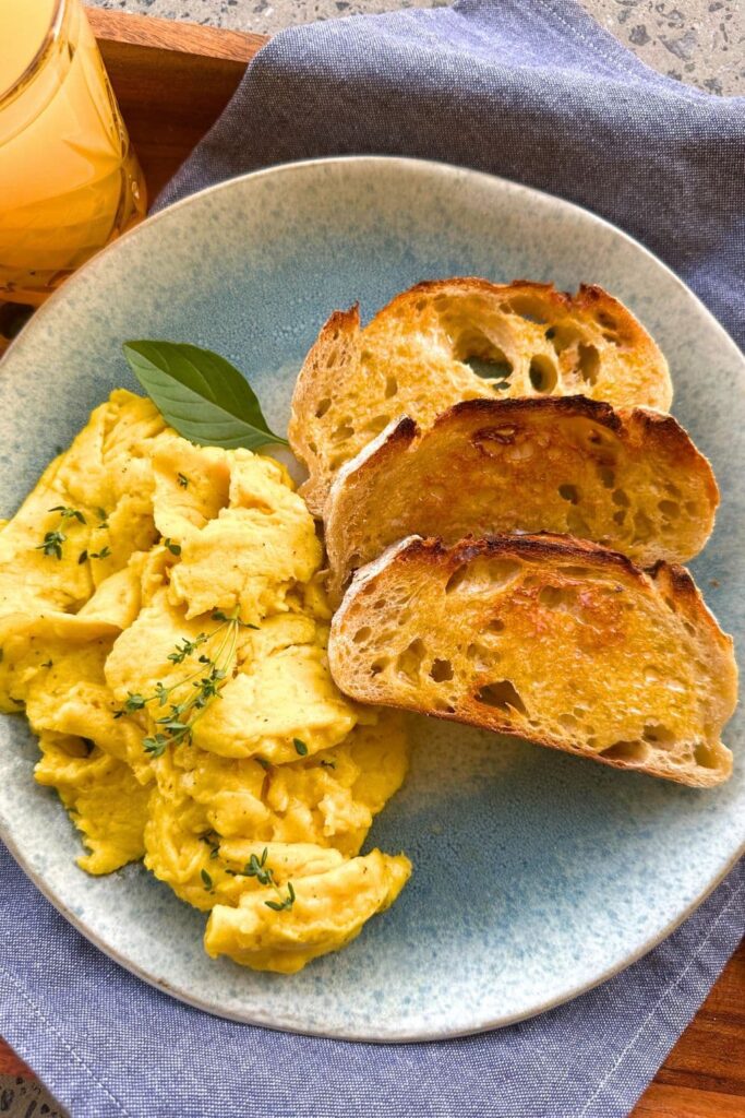 A blue stoneware plate topped with sourdough scrambled eggs, 3 slices of buttered sourdough toast and sitting on a blue dish towel. There is also a glass of orange juice to the left of the plate.