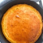 A cast-iron skillet containing sourdough cornbread that has just come out of the oven. It is golden brown and surrounded by a blue dish towel.