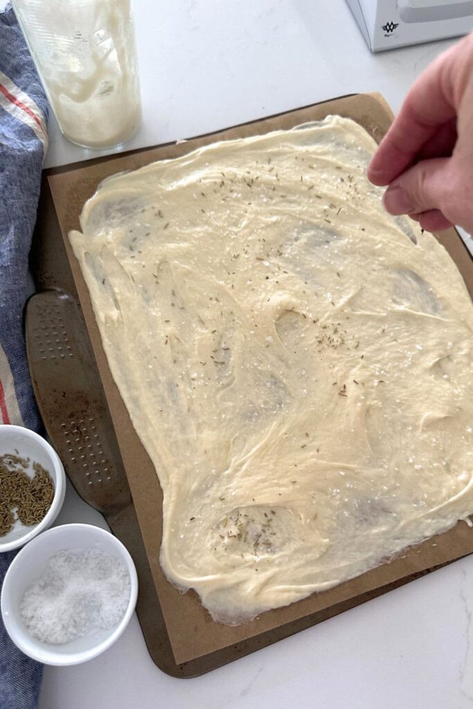 A baking tray spread with sourdough discard cracker mixture. You can see the small bowls of rosemary and salt used to season the crackers.